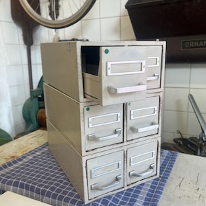 Puede incluir: Una pila de seis cajones de archivo de metal vintage en color beige claro. Cada cajón tiene un asa plateada y un portaetiquetas. Un cajón está abierto, revelando su interior. Los cajones están apilados sobre un paño a cuadros azul y blanco.