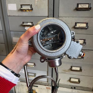 May include: A silver-colored industrial gauge with a clear, circular face displaying internal components and numerical markings. A coiled metal tube and a straight metal probe extend from the gauge's body. A red light is visible on the side.