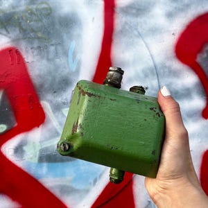 Może przedstawiać: Zwietrzały, zielony, prostokątny metalowy przedmiot z trzema cylindrycznymi mocowaniami. Obiekt jest trzymany na tle graffiti z czerwoną i białą farbą.