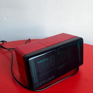 May include: A retro red and black digital alarm clock with a rectangular shape. The clock shows the time in green digits and has the text "Электроника 73". The clock is on a red surface.