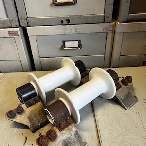 May include: Two white ceramic insulators with black and brown components, resting on a weathered surface. The insulators have a cylindrical shape with a flared top and base, and are attached to metal brackets.