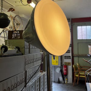 以下が含まれることがあります： 金属製のポールと幅広のオフホワイトのシェードを備えた、大きな円錐形のフロアランプ。 ランプはオリーブグリーンで、温かみのある光を放ちます。 背景には、ファイルキャビネットやヴィンテージの装飾が含まれています。