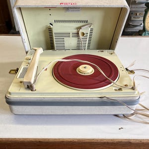 Op de afbeelding: Vintage draagbare platenspeler met een crèmekleurige buitenkant en een rode vinylplaat. De speler heeft een scharnierend deksel en een ingebouwde luidspreker. Het ontwerp is retro, met de nadruk op functionaliteit en draagbaarheid. De speler is open en klaar om te spelen.