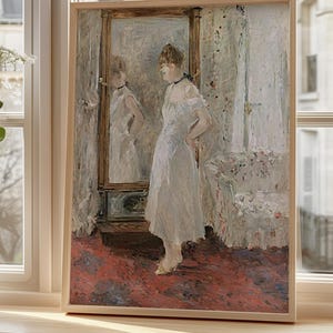 Puede incluir: Una impresión enmarcada de una mujer con un vestido blanco frente a un espejo. La obra de arte tiene un estilo suave e impresionista con tonos apagados de blanco, rojo y beige. La impresión se exhibe en un alféizar de madera.