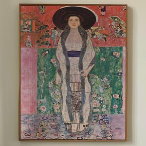 May include: A framed print of a Gustav Klimt painting. The artwork depicts a woman in a long, patterned dress and a large hat. The background features floral and geometric patterns in shades of red, green, and pink. The print is displayed above a bed.