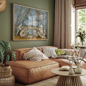 May include: A living room scene featuring a large framed artwork depicting figures near water, above a brown leather sectional sofa. Decorative pillows in white, black, and green adorn the sofa. A round wooden coffee table and potted plants complete the cozy setting.