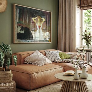 May include: A framed print of a painting hangs above a brown leather sectional sofa. The painting depicts a woman in a white dress. A round wooden coffee table with two white vases and a book sits in front of the sofa. A potted plant is on the left.