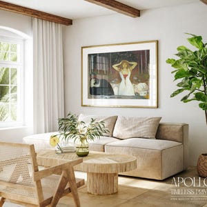 May include: A living room scene featuring a framed artwork with a gold frame, a beige sectional sofa, and a wooden coffee table. A large green plant in a woven basket sits in the corner. The room has a neutral color palette with natural light.