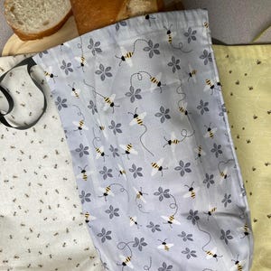 May include: Three fabric bread bags with drawstring closures. One bag is light gray with a pattern of bees and gray flowers, one is cream with tiny brown bees, and one is yellow with a delicate bee and swirling line pattern.