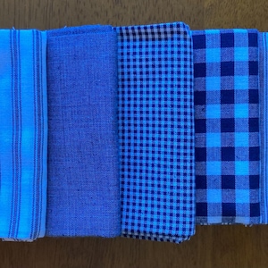 May include: Five folded fabric napkins arranged in a row. The set includes a white napkin with thin blue stripes, a solid blue napkin, a small blue and white gingham napkin, a larger blue and white checkered napkin, and a blue striped napkin.