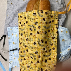 Puede incluir: Una barra de pan se encuentra dentro de una bolsa de pan amarilla y blanca con temática de abejas. La bolsa está abierta, revelando el pan. La bolsa está rodeada de otras bolsas de tela con estampados florales y de abejas. Las bolsas tienen cordones y son de tela.