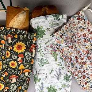 May include: Three fabric bread bags with drawstring closures. The left bag features a dark green background with yellow sunflowers and orange mushrooms. The center bag is white with green herb illustrations and text including CHIVES. The right bag has a white background with a dense pattern of small red, pink, and blue flowers.