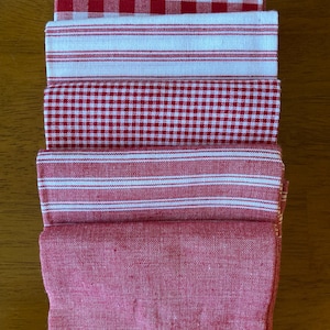 May include: A stack of five folded fabric napkins in various red and white patterns. The collection includes a large red and white gingham check, a white napkin with thin red stripes, a small red and white gingham check, a red napkin with white stripes, and a solid red napkin.