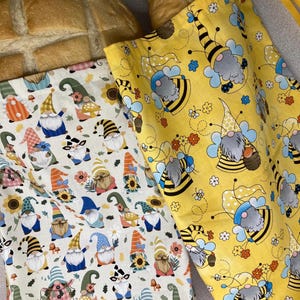 May include: Two fabric bread bags with colorful gnome and bee patterns. One bag is yellow with bee and gnome designs, the other is off-white with various gnome designs. Both bags have a drawstring closure and are filled with bread.