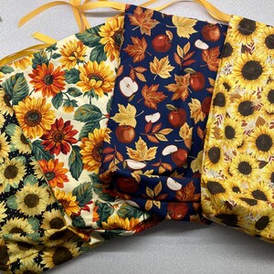 May include: Four fabric drawstring bags featuring distinct patterns: a black background with sunflowers, a cream background with sunflowers and green leaves, a navy blue background with autumn leaves and red apples, and a cream background with sunflowers.