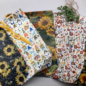 May include: A collection of five fabric drawstring bags featuring various patterns including yellow sunflowers on black, colorful gnomes on white, large sunflowers on green, small floral prints on white, and mushrooms with sunflowers on dark green.