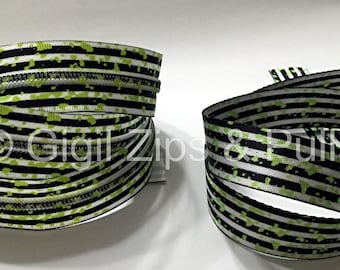 Op de afbeelding: Twee spoelen lint met een gestreept patroon in zwart, wit en groen. Het lint heeft een herhalend patroon van zwarte strepen en groene spetters. De tekst "Gigi Zips & Puffs" is zichtbaar op het lint.