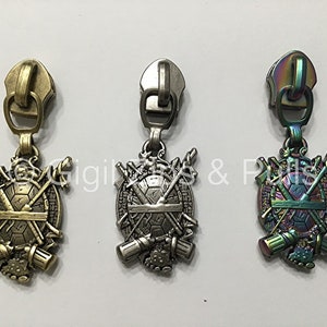 May include: Three zipper pulls with a turtle and weapons design. The pulls are in gold, silver, and iridescent colors.
