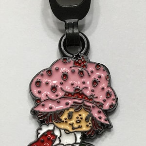 May include: A metal zipper pull featuring a cartoon character in a pink bonnet with red strawberry accents. The character is wearing a red and white dress, green pants, and brown shoes. The zipper pull is attached to a black metal zipper.