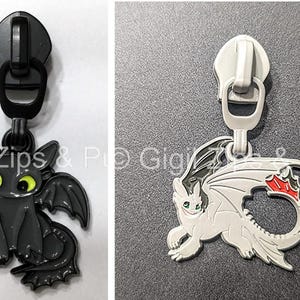 Puede incluir: Dos tiradores de cremallera con diseños de dragones. Uno es negro con ojos amarillos, el otro es blanco con detalles rojos y negros. Ambos tienen tiradores de cremallera metálicos.