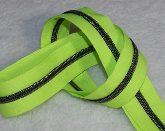 Op de afbeelding: Een neon groene rits met iriserende tanden, in lussen gelegd op een witte ondergrond. De ritsband is felgroen en de tanden hebben een metallic, veelkleurige glans. Geschikt voor diverse naaiprojecten.