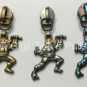 May include: Three zipper pulls featuring a cartoon Teenage Mutant Ninja Turtle in a fighting pose. The zipper pulls are in three different finishes: gold, silver, and iridescent.