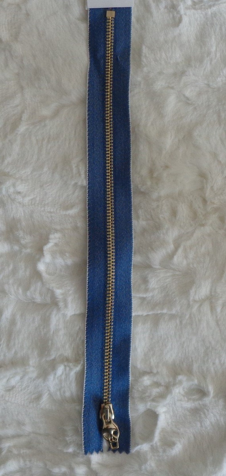 May include: A gold zipper with a blue denim tape. The zipper pull is a gold skull.