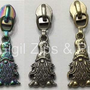 May include: Three zipper pulls with a gnome design. The gnome is wearing a hat and has a beard. The zipper pulls are available in iridescent, silver, and gold.