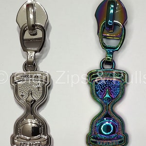 May include: Two zipper pulls with an hourglass design. The hourglass is filled with stars and a rocket ship. The text "TIME TO" and "EXPLORE" is on the hourglass. One pull is silver and the other is iridescent.