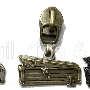 May include: Three metal zipper pulls with coffin-shaped charms. The charms feature a skeleton hand reaching out from the coffin. The zipper pulls are silver, bronze, and black.