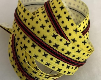 Op de afbeelding: Een opgerolde ritsband met een felgele achtergrond en een herhalend patroon van kleine zwarte kruisjes en stippen. De rits is zwart met een dunne rode accentlijn die langs het midden van de band loopt voor een opvallend contrast.