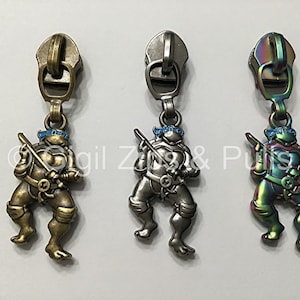 May include: Three zipper pulls featuring a Teenage Mutant Ninja Turtles character. The pulls are in bronze, silver, and iridescent finishes.