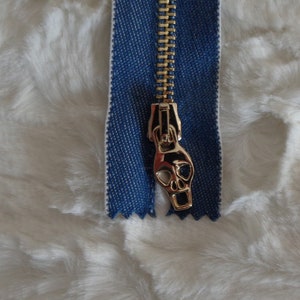 May include: A gold zipper with a skull-shaped pull. The zipper is attached to a blue denim fabric.