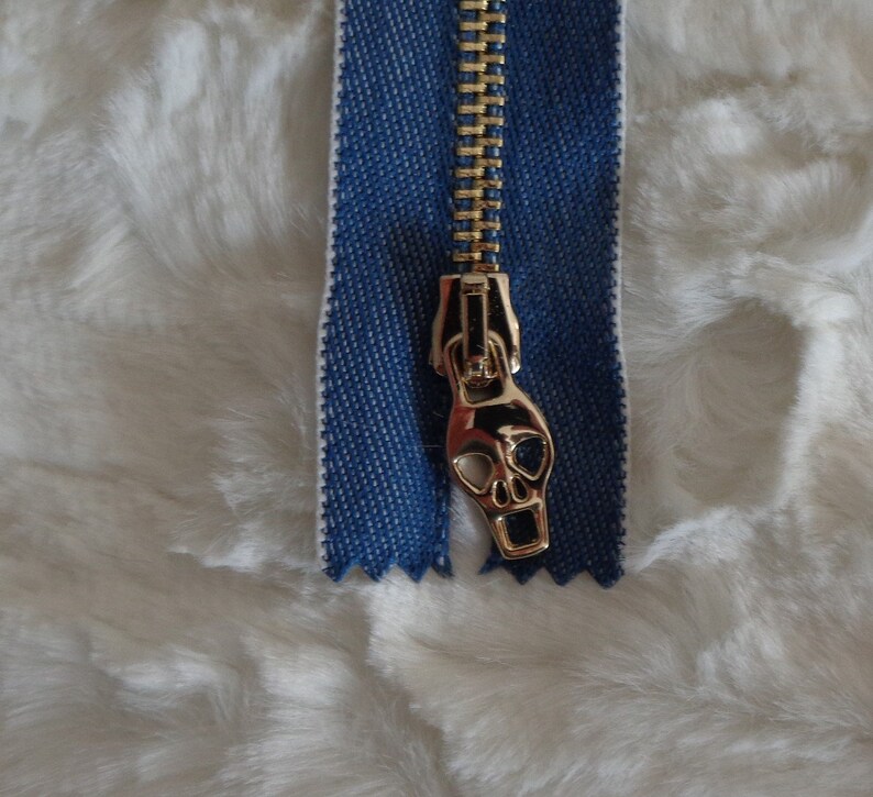 May include: A gold zipper with a skull-shaped pull. The zipper is attached to a blue denim fabric.