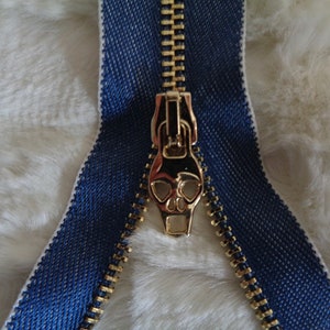 May include: A gold zipper with a skull-shaped pull. The zipper is sewn into a blue fabric tape.