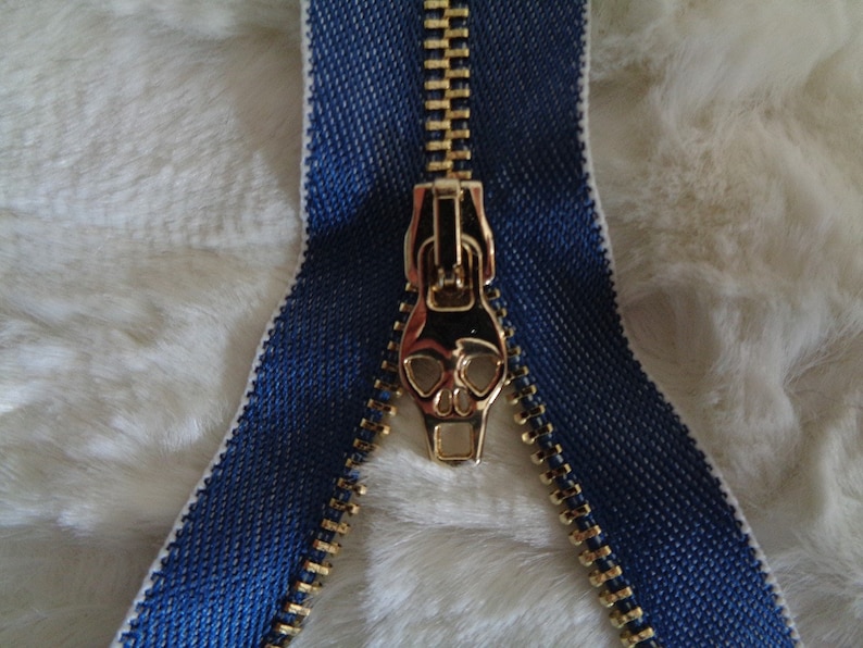 May include: A gold zipper with a skull-shaped pull. The zipper is sewn into a blue fabric tape.