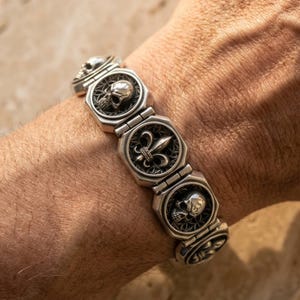 Puede incluir: Pulsera plateada con diseños de calaveras y flor de lis. La pulsera está compuesta por múltiples eslabones octagonales, cada uno con un emblema detallado. Las calaveras y la flor de lis están dentro de marcos circulares.