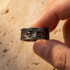 Könnte beinhalten: Ein silberfarbener Ring mit einem detaillierten Design von zwei Augen. Der Ring hat ein strukturiertes Band mit einem eingravierten Gesicht. Der Ring wird zwischen den Fingern gehalten und zeigt die aufwendige Handwerkskunst.