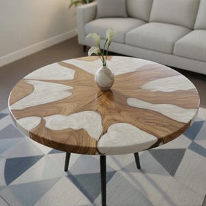 Puede incluir: Mesa de centro redonda con tapa de madera y resina blanca. La mesa tiene un diseño de vetas de madera natural con detalles de resina blanca. Se apoya sobre tres patas de metal negro. Un pequeño jarrón blanco con flores está sobre la mesa.