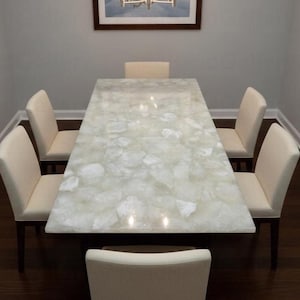 May include: A rectangular dining table with a light-colored, patterned surface, surrounded by six cream-colored chairs. The table is the focal point, with a unique, textured design. The chairs have dark wooden legs. A framed picture hangs on the wall.