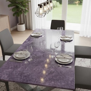 Puede incluir: Una mesa de comedor con una superficie morada texturizada está preparada para una comida. La mesa está rodeada de sillas grises y está puesta con platos blancos, cubiertos y copas de vino. Una moderna lámpara cuelga sobre la mesa.