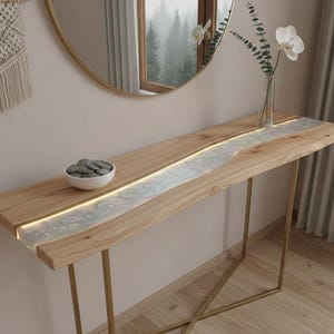 Puede incluir: Una consola de madera con una tapa de madera clara y una incrustación central de resina nacarada e iluminada. La mesa tiene patas de metal dorado y un pequeño cuenco blanco con piedras grises. Un espejo decorativo y flores están en el fondo.