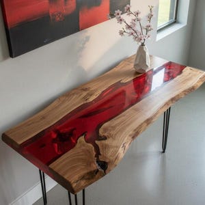 May include: A live edge wooden table with a vibrant red resin river. The table has black hairpin legs and a natural wood grain finish. A white vase with pink flowers sits on the table. The table measures approximately 60 inches long.