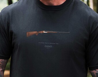 May include: Black t-shirt featuring a rifle graphic with brown and silver details. The text "SILENCE HAS A TRIGGER TOO" and "OHNO NYC" are printed below the rifle. The shirt is made of a soft, dark fabric.