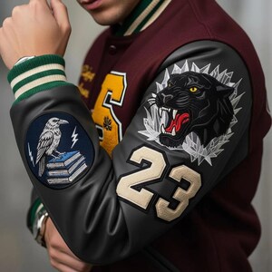 Könnte beinhalten: Burgunderrote und schwarze Collegejacke mit einem schwarzen Panther-Aufnäher auf dem Ärmel. Die Jacke hat einen grün-weiß gestreiften Kragen und Bündchen. Der linke Ärmel hat einen Raben- und Buch-Aufnäher, der rechte die Nummer 23.