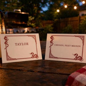Könnte beinhalten: Zwei cremefarbene Tischkarten mit roten Drachen- und Bordürendesigns. Auf einer Karte steht "TAYLOR" und auf der anderen "CHICKEN, FILET MIGNON". Die Karten liegen auf einem Holztisch, mit Laternen und einem Grill im Hintergrund.