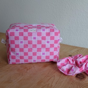 May include: A pink and white checkered cosmetic bag and matching scrunchie. The bag has a zipper closure and white handles. The bag and scrunchie are decorated with pink starbursts. The bag has the initials "MM" on a white label.