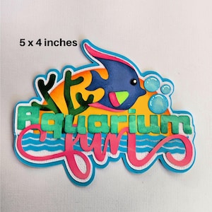May include: A colorful paper craft with the words "Aquarium Fun" in a whimsical font. The craft features a blue fish with green and yellow accents, coral, and bubbles. The craft is 5 inches by 4 inches.