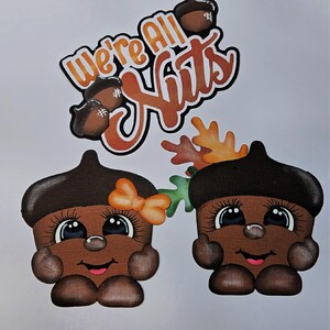 May include: A whimsical fall-themed design featuring two cartoon acorns with smiling faces, eyelashes, and bow accents. The text "We're All Nuts" is displayed above the acorns in orange and brown colors. The acorns are brown with dark brown caps.