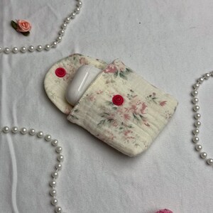 Könnte beinhalten: Ein AirPods-Etui mit floralem Muster und passendem Halter mit roten Knopfverschlüssen. Das Etui ist cremefarben mit rosa und grünen Blumenmustern. Ein Paar weiße AirPods befinden sich im Inneren. Dekorative Perlen und eine rosa Blume im Hintergrund.